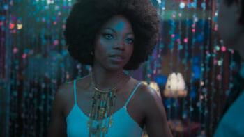 Paola González wears a white tank top and an intricate gold necklace. She stands in front of a beaded curtain.