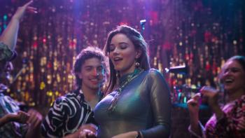 Laura Taylor wears a sheer blue shirt and blue top underneath, with gold accessories. She dances in a nightclub.