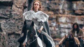 The Winter King TV show screenshot: A regal looking woman rides a horse wearing a fur shawl in front of a castle.