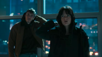 The Kollective screenshot: A young White man and a young White woman (both dark-haired) take of their hoods in front of a window in a tall building