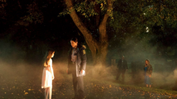 A female officer stand in front of a girl in a white dress at night in front of a big tree with a small crowd beneath it