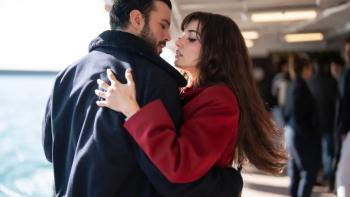 A brunette woman in a red coat embraces a dark-haired man in a black leather jacket in an airport-like setting