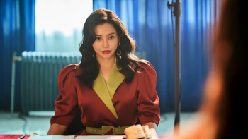 An Asian woman with curled hair in a leather blazer sits in what looks like a conference room and stars intensely at someone across the table