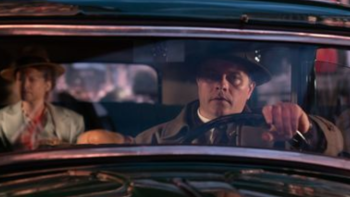 A detective-looking White man with a hat is driving a car with a blurred woman in the backseat