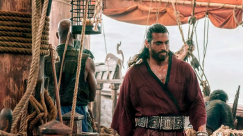 A long-haired man in a dark red tunic walks across a wooden ship deck surrounded by ropes, rigging, and crew members.