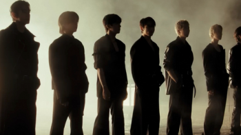 A group of people stand in a line as dark silhouettes, backlit by a bright, hazy light, creating a dramatic and shadowy scene.