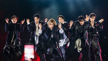 A group of performers move in sync on a brightly lit stage, captured mid-choreography with expressive hand gestures, while a crowd watches from below.
