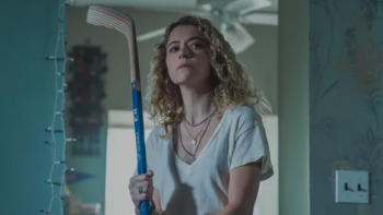Curly-haired woman holding a hockey stick indoors, looking determined.