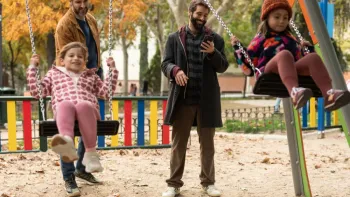 Shared Custody TV: Diego and a male friend stand behind their daughters on the swing in a park during Fall. Diego looks at this phone and smiles.