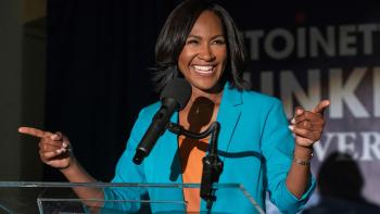 Terri J. Vaughn stands at a podium, pointing her fingers out to the side. She wears a blue suit jacket and orange shirt.