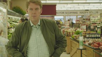 A man with red hair and an olive green jacket stands in a grocery store with a serious expression on his face.