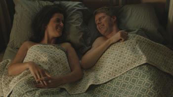 A woman and man are pictured laying in bed, under a blue and white patterned blanket, smiling at one another.