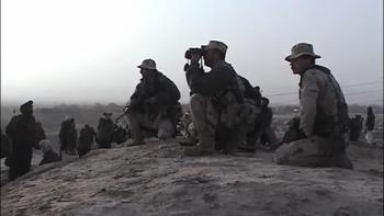 Armed soldiers sit on the ground outside on a dirt hill. One soldier holds up a pair of binoculars.