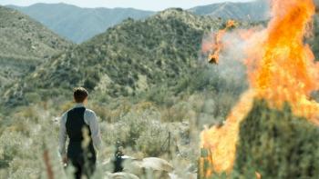 Strange Angel TV Series: A man in 1930s-period clothing stands by desolate mountains, watching a rocket take off in a fiery explosion. 