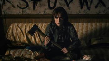Stranger Things TV show screenshot 2: Joyce sits on her couch clutching an axe with an alphabet painted on the wall behind her
