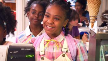 Stranger Things 3 TV show screenshot 3: Erica smiles in front of a cash register, dressed in a pink 1980s dress