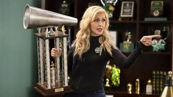 Stumble TV show screenshot: A White, blonde woman stands in an office holding a huge cheerleading trophy