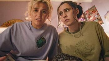 Such Brave Girls TV show screenshot 3:Lizzie Davidson stares at the screen while Kat Sadler side eyes her. They sit on a bed.