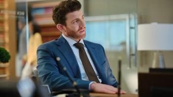 Bryan Greenberg sits at a desk. He wears a blue suit jacket, light blue shirt, and brown tie.