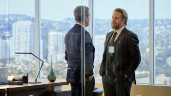 Stephen Amell and Josh McDermitt stand in front of each other in an office with large windows showing a backdrop of L.A. 