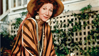 Tales of the City TV Show: A sixtysomething woman in bohemian clothing and a straw hat works in a garden in front of a leafy trellis. 