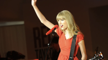 Taylor TV show screenshot 2: Taylor Swift in orange dress and guitar on stage with arn held up.