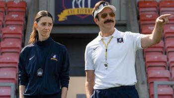 Ted Lasso Season 4: Ted gestures at something on the football pitch as Alice stands beside him, looking irritated; both wear manager uniforms