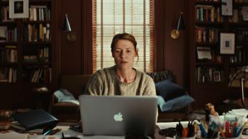Claire Danes sits at a cluttered desk, working on a silver MacBook laptop. She wears a grey sweater.