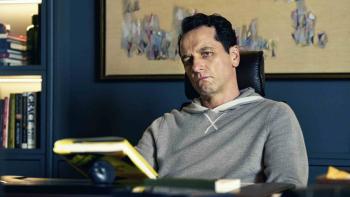 Matthew Rhys sits in a chair at minimal desk. He holds a hardback book. He wears a gray hooded shirt.