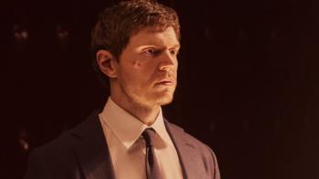 Ashton Kutcher stands in front of a helicopter. He wears a dark suit jacket, dark shirt, and brown sunglasses.