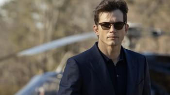 Ashton Kutcher stands in front of a helicopter. He wears a dark suit jacket, dark shirt, and brown sunglasses.