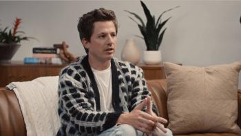 The Charlie Puth Show TV Show screenshot 3: Puth wears a black and white striped cardigan. He sits on a born leather couch in an office.