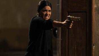 The Dead Girls TV show screenshot 1: Paulina Gaitán stands in the middle of open doors. She aims a handgun. She's dressed in black. 