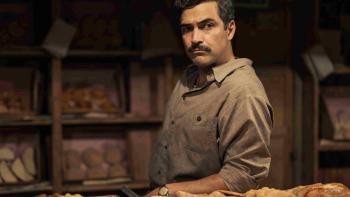 The Dead Girls TV show screenshot 3: Alfonso Herrera stands behind trays full of pastries. He wears a tan button-up shirt and a watch.