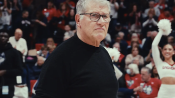 The Dynasty: UConn Huskies TV show screenshot 1: Coach Geno Auriemma in a black sweater stands on a basketball court, surrounded by cheering fans.