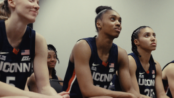 The Dynasty: UConn Huskies TV show screenshot 2: Paige Beuckers and teammatesin navy jerseys sitting, attentively listening.