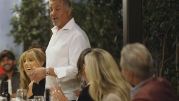The Family Stallone screenshot 2: Sylvestor Stallone in white shirt stands and talks to table of dinner guests. 