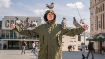 The Full Monty TV show: Gaz stands in a city plaza in an olive colored military jacket. The hood of the jacket is up and his arms are outstretched. Balanced on his arms and on top of his head are six pigeons, who appear to be peacefully resting. 