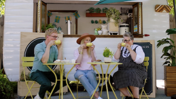 The Gentle Art of Swedish Death Cleaning screenshot 2: Johan, Kat, and Ella sip yellow coffee cups on yellow chairs in front of Frika stand.