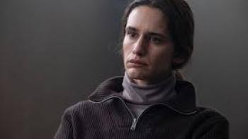 Valentina Belle in a dark pullover sweater and a turtleneck, sitting in a gray room.