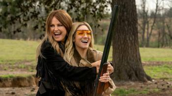 The Hunting Wives TV show screenshot 1: Margo holds Sophie in her arms while Sophie points a rifle into the air; both are in a woodsy area