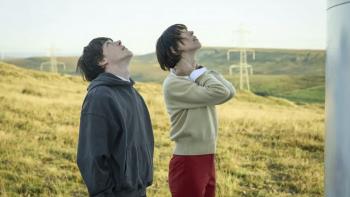 The Listeners TV show screenshot 2: Ollie West and Rebecca Hall stand in a field. West wears a dark hoodie; Hall wears a yellow sweater and red pants.