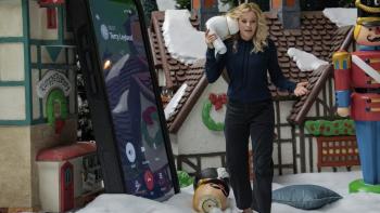 The Miniature Wife TV show screenshot 3: Banks holds a giant ear bud. She stands next to a giant smart phone. She wears a blue shirt and black pants. 
