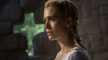 The Pendragon Cycle: Rise of the Merlin TV show screenshot 2: Charis in white medieval outfit with braided hair, standing in a dimly lit stone room.