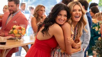 Eve Hewson in a red dress hugs Meghann Fahy from behind as they dance and laugh together. People dance and drink around them.