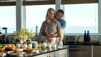 Liev Schreiber hugs Nicole Kidman while kissing her neck. They stand in a luxurious kitchen and the ocean in the background.