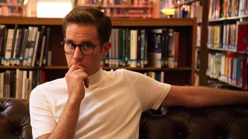 The Politician TV Series: Payton puts his hand to his chin and looks thoughtful in front of library shelves lined with books. 