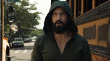 The Punisher: One Last Kill TV show screenshot 3: Jon Bernthal walks in between two yellow school buses. He wears a green zip up hoodie.