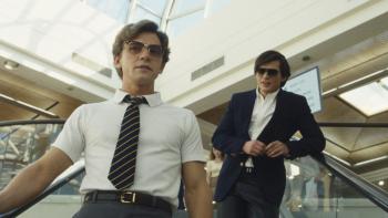Igby Rigney and Homer Gere stand on a escalator in a mall. They both wear dark blue and white school uniforms, and sunglasses.