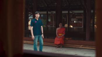 The Believers TV show screenshot: A young Thai man is shown standing in a temple near a sitting monk.